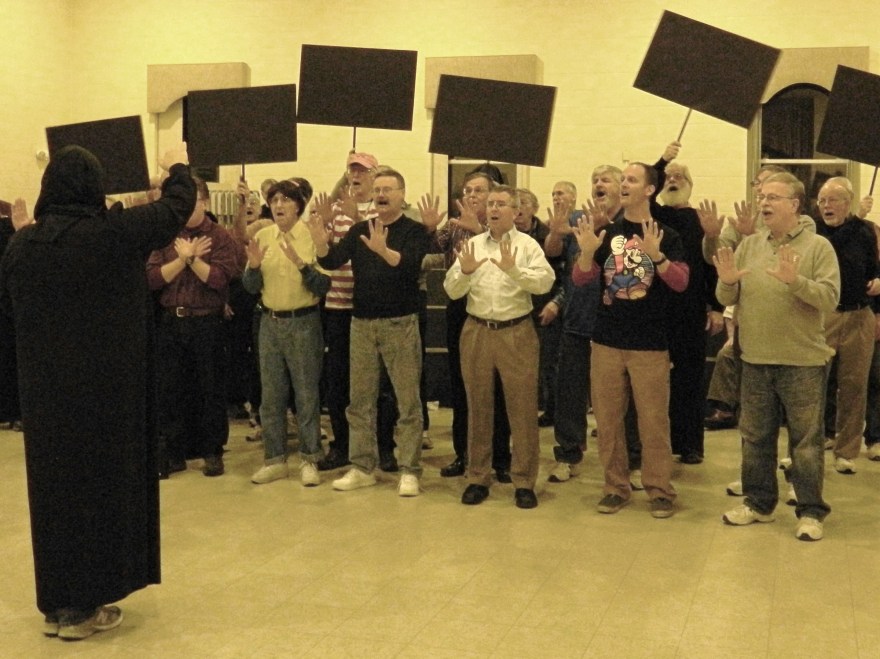 A black light will used for this number so all the audience will see is the motion of hands in white - thus, the chorus director is shroud in a black. 
